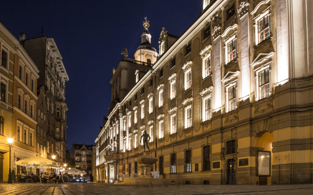 Oświetlony barokowy gmach Uniwersytetu Wrocławskiego nocą z Fontanną Szermierza na środku dziedzińca.
