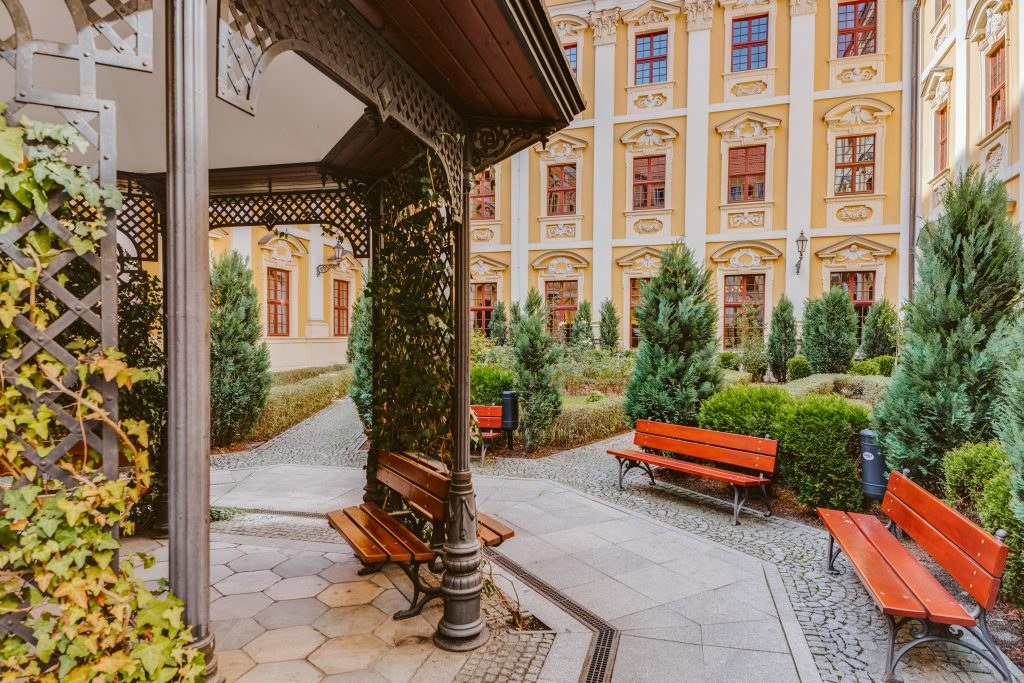 Zadbany ogród z czerwonymi ławkami i ozdobnym żelaznym zadaszeniem przy budynku.